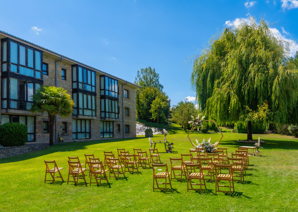 Chairs set out at a wedding venue next to a large building and a willow tree.
